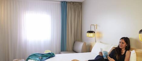 Una habitación de hotel con cama, lámpara de lectura, ventana con cortinas y mesita de noche.