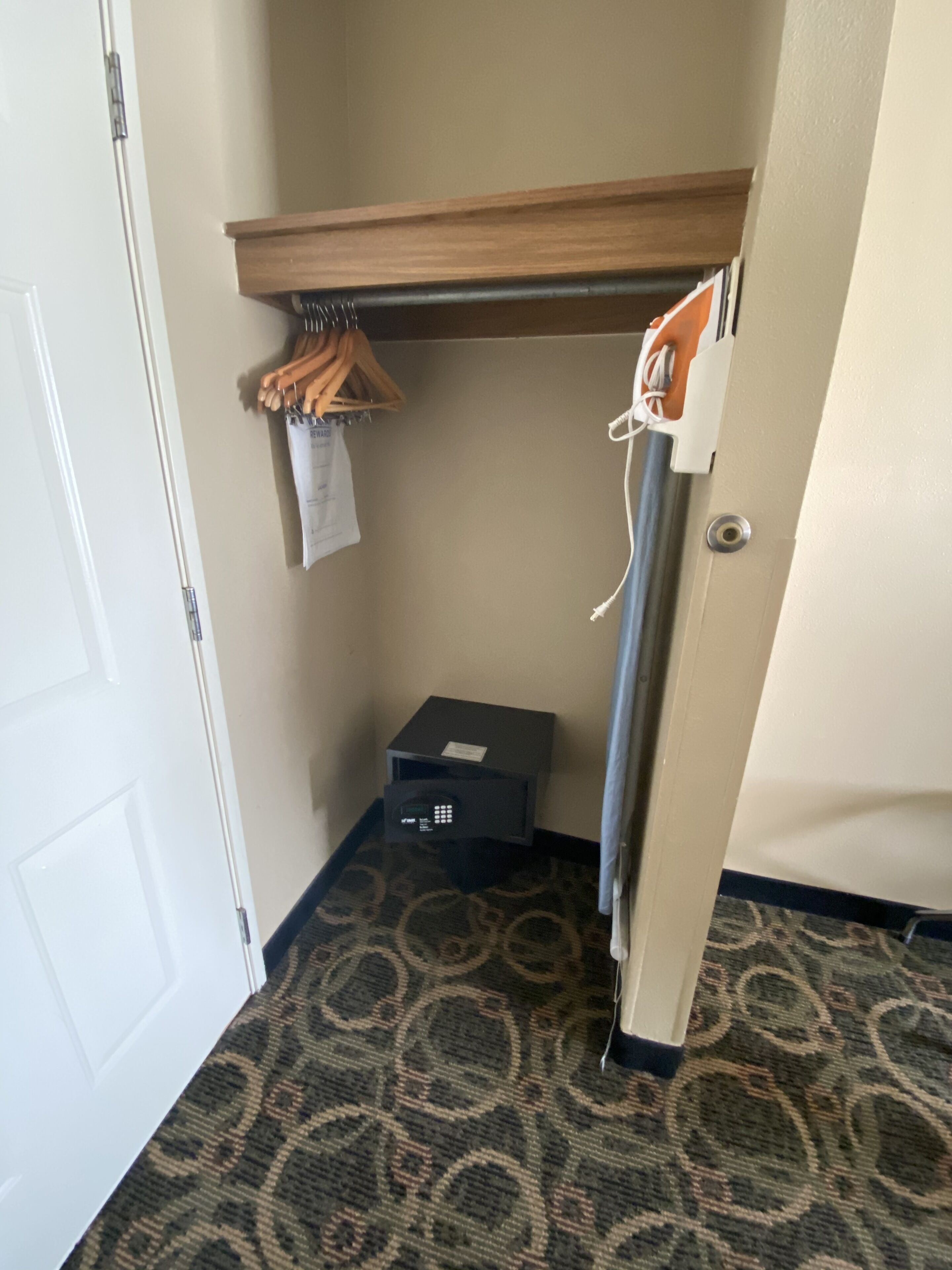 A hotel room with a small closet, a coat rack with a coat, a suitcase, and a small table with a remote control.