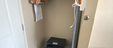 A hotel room with a small closet, a coat rack with a coat, a suitcase, and a small table with a remote control.
