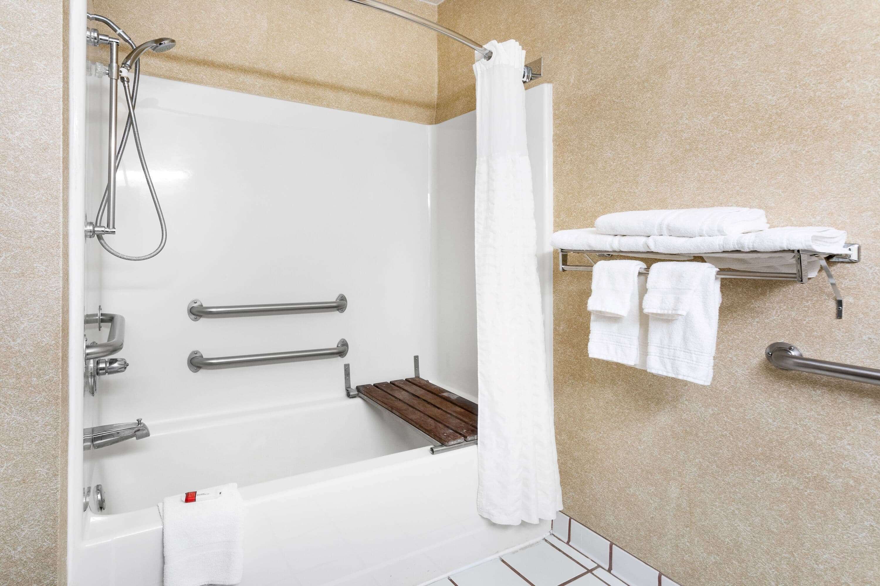 A modern bathroom with a bathtub, shower, and handrails. There are towels and a shelf with more towels.