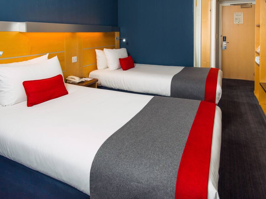 Two adjacent hotel beds with white bedding, red and grey accents, a bedside table with a lamp, and a wooden headboard.