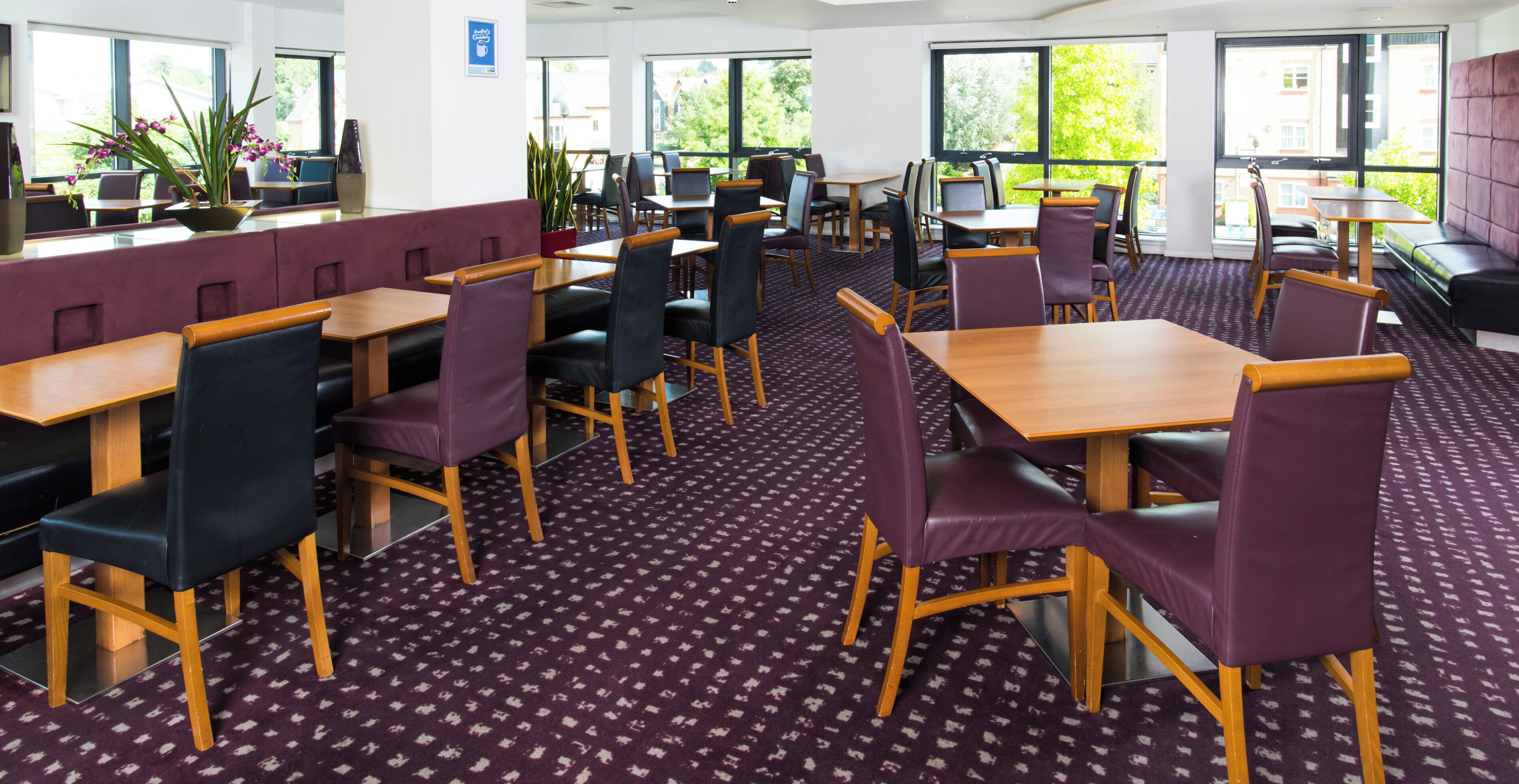 A modern dining area with multiple tables and chairs, large windows with a view of greenery, and a patterned carpet.