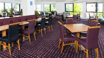 A modern dining area with multiple tables and chairs, large windows with a view of greenery, and a patterned carpet.