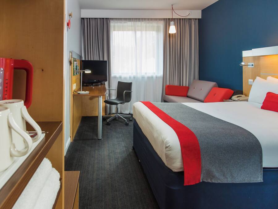 A hotel room with a bed, desk, chair, television, and a red telephone.