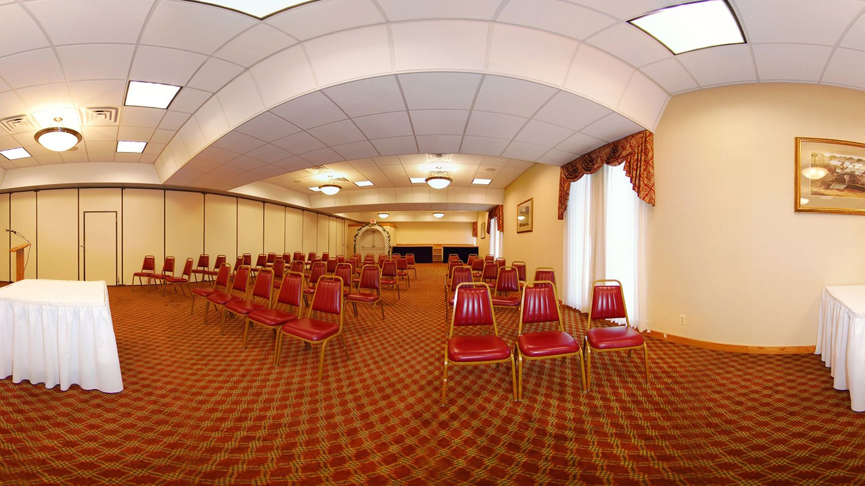 Uma sala de conferências com fileiras de cadeiras vermelhas, uma mesa branca e um tapete estampado.