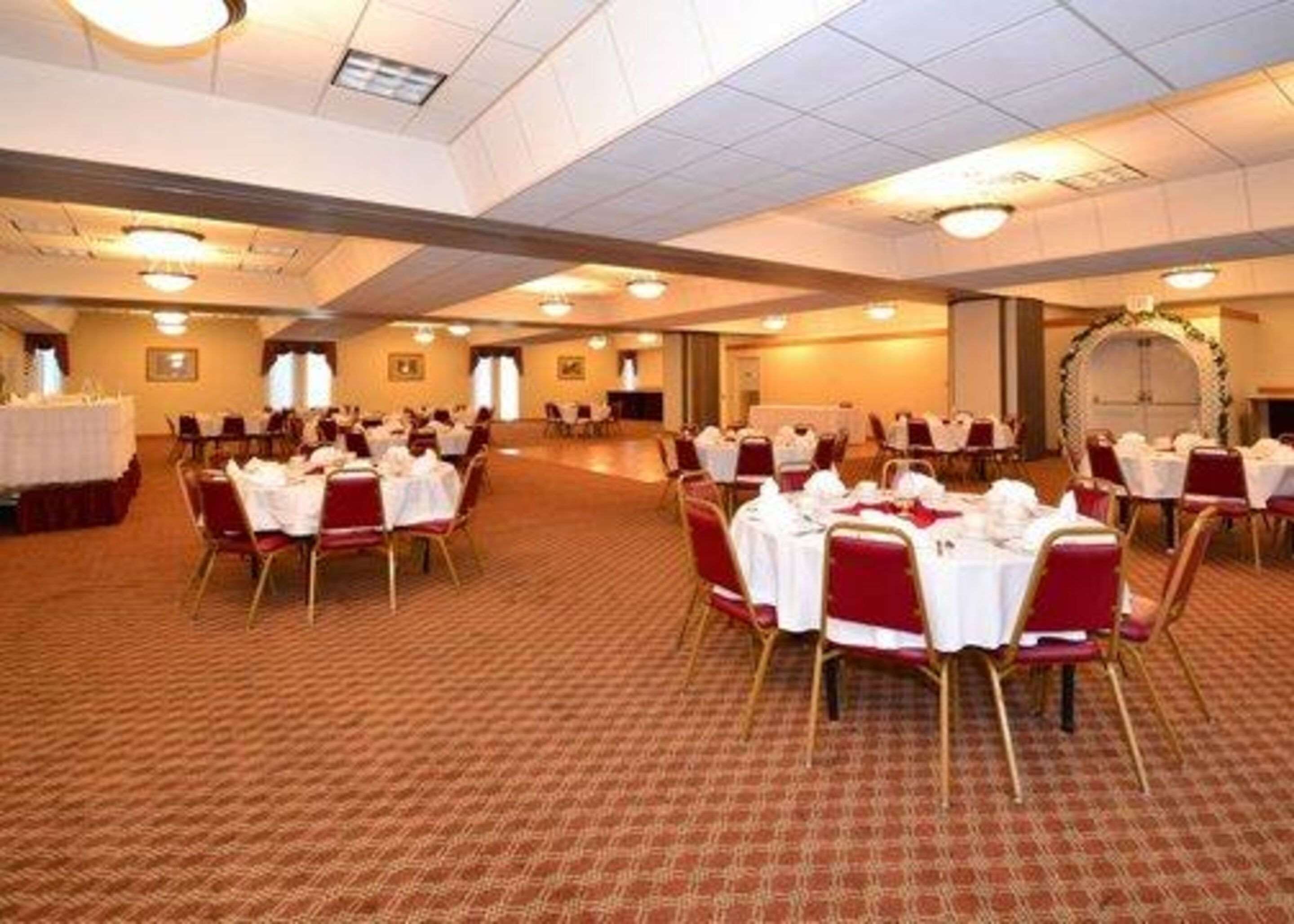 A large banquet hall with round tables set for dining, carpeted flooring, and a high ceiling with recessed lighting.