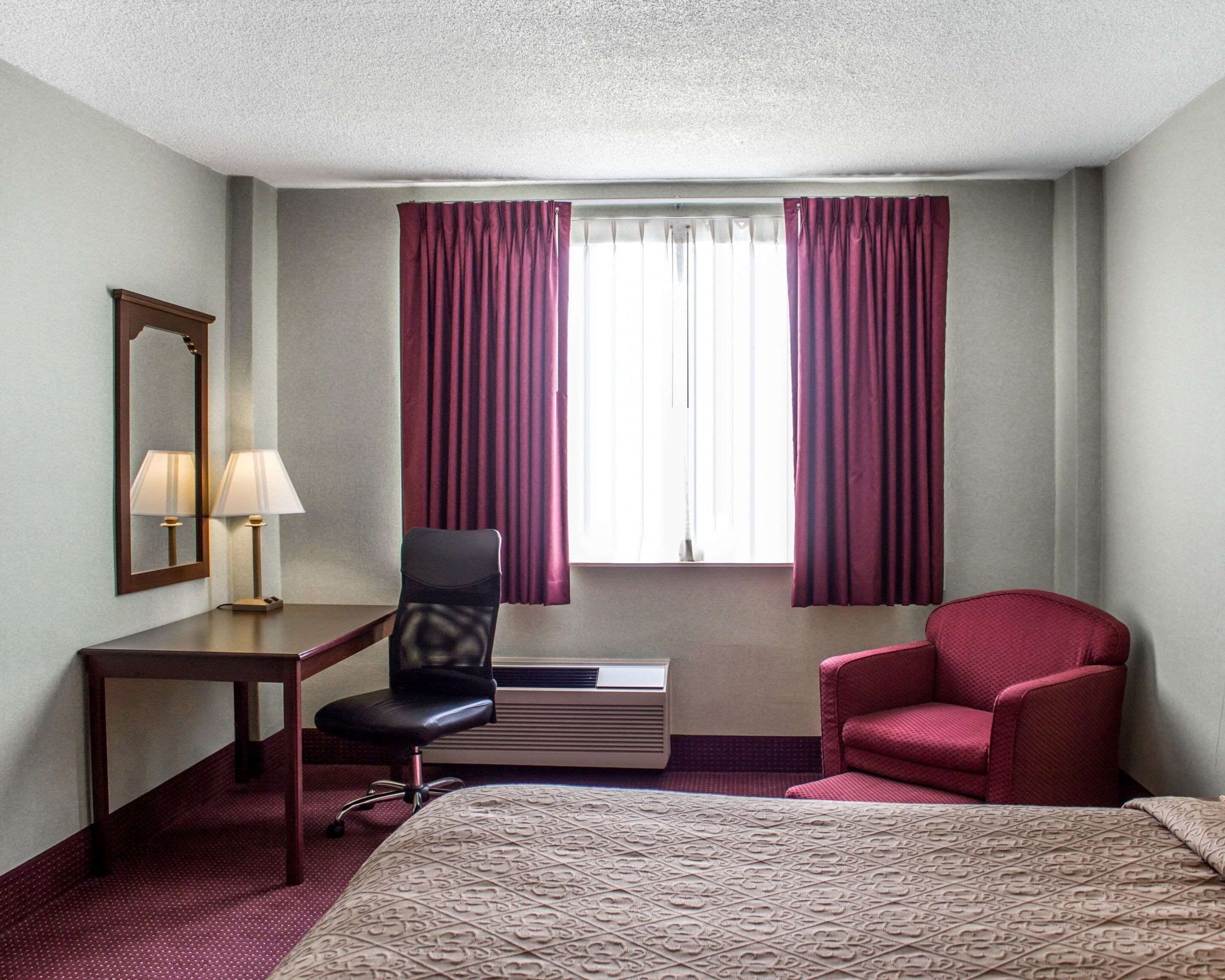 A hotel room with a bed, a desk with a chair, a mirror, and two windows with curtains.