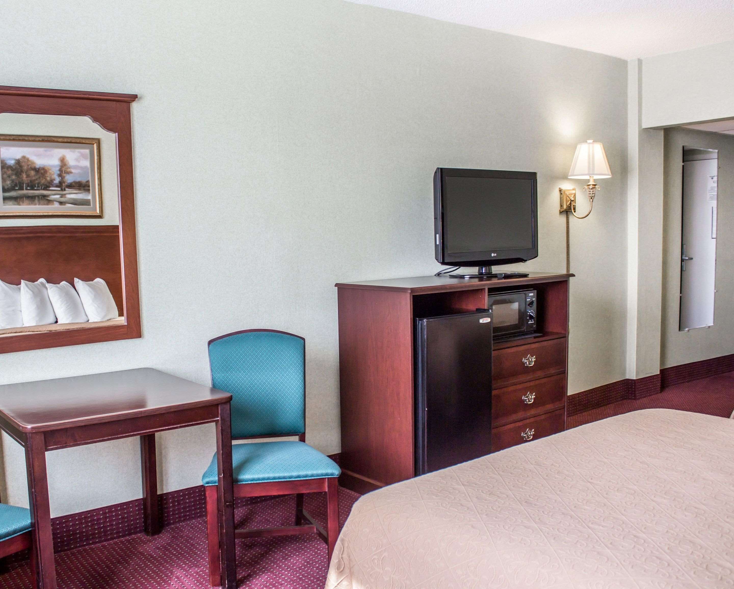 A hotel room with a bed, a desk, a chair, a television, a microwave, a refrigerator, and a wall-mounted lamp.