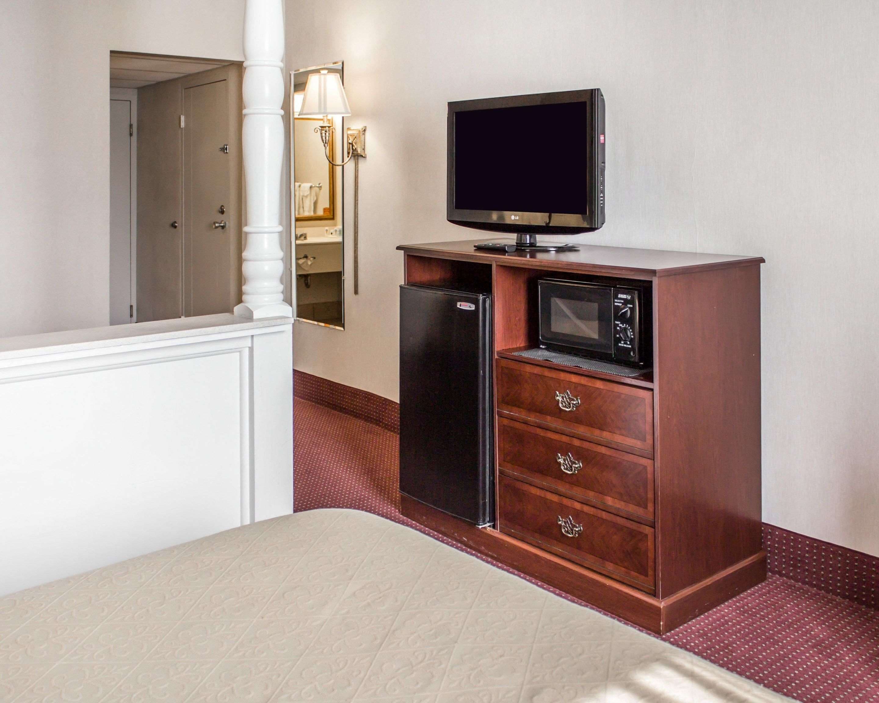 A hotel room with a bed, a minibar with a TV and microwave, a mirror, and a wall-mounted light fixture.