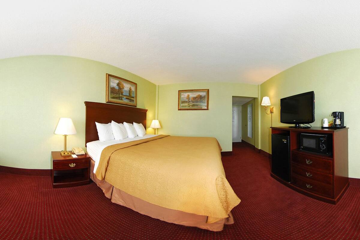 A hotel room with a bed, two bedside tables with lamps, a red armchair, a television, and a minibar.