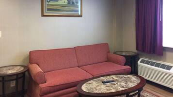 Una sala de estar con un sofá rojo, dos mesitas auxiliares, una mesa de centro redonda, un cuadro enmarco de un paisaje, una ventana con cortinas moradas y un aire acondicionado de pared.