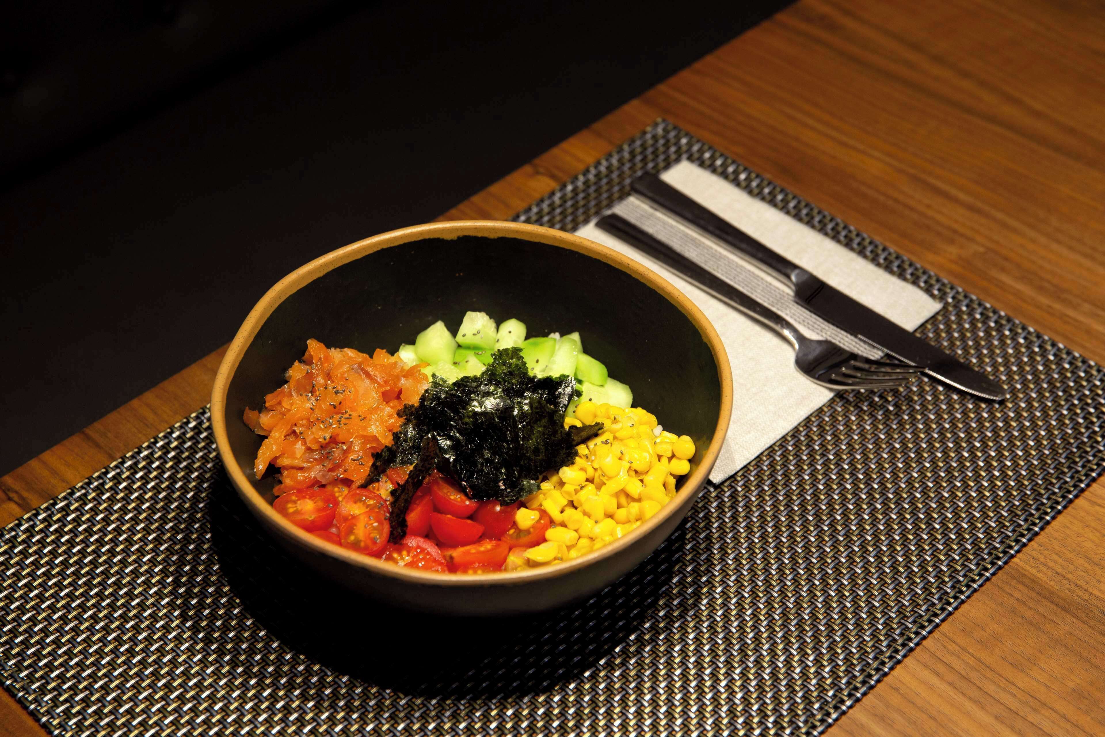 A bowl of food on a black plate with a fork and knife on a woven placemat.