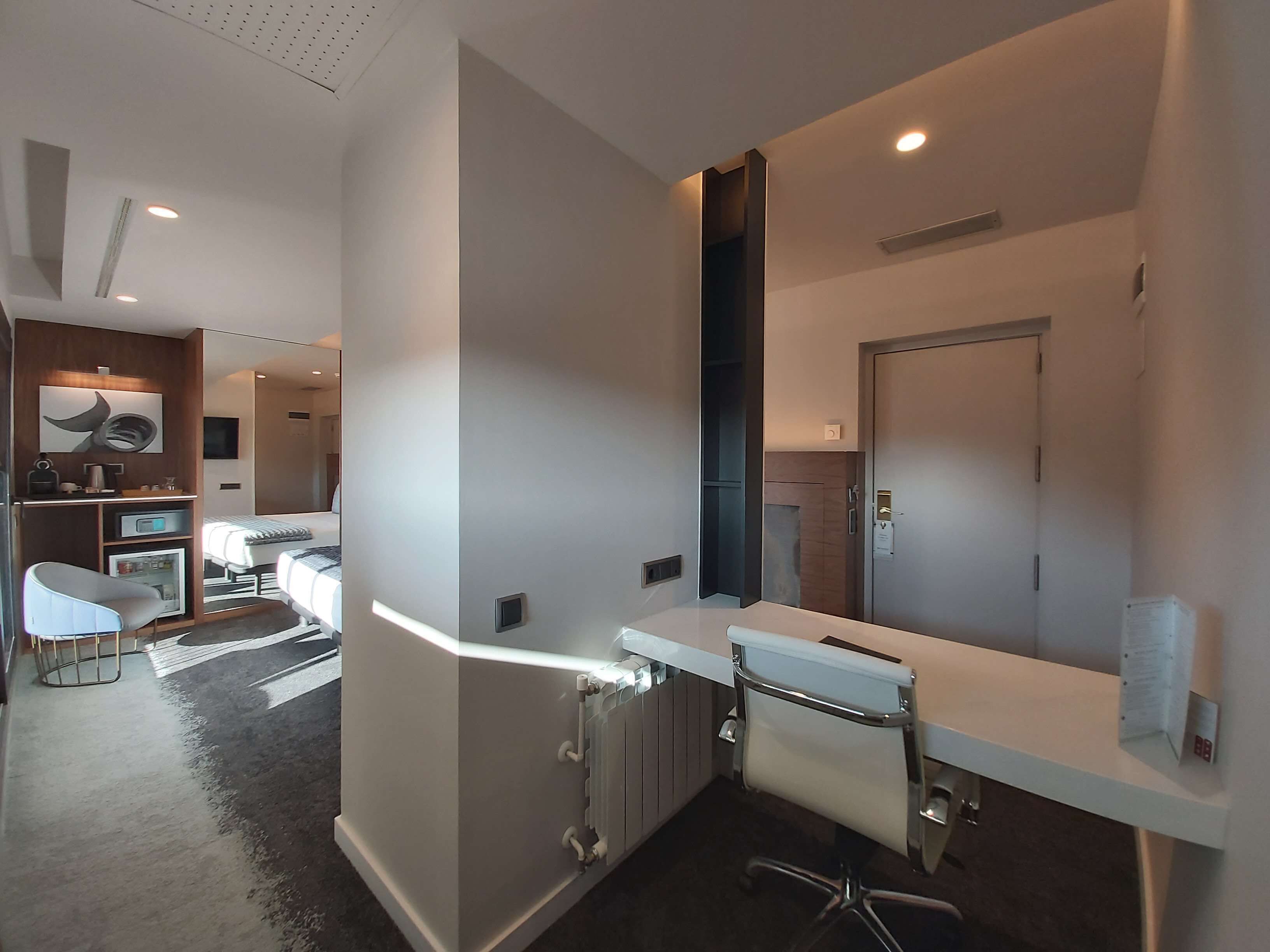 A modern hotel room with a large bed, a desk, and a chair. There is a bathroom with a toilet and a shower.