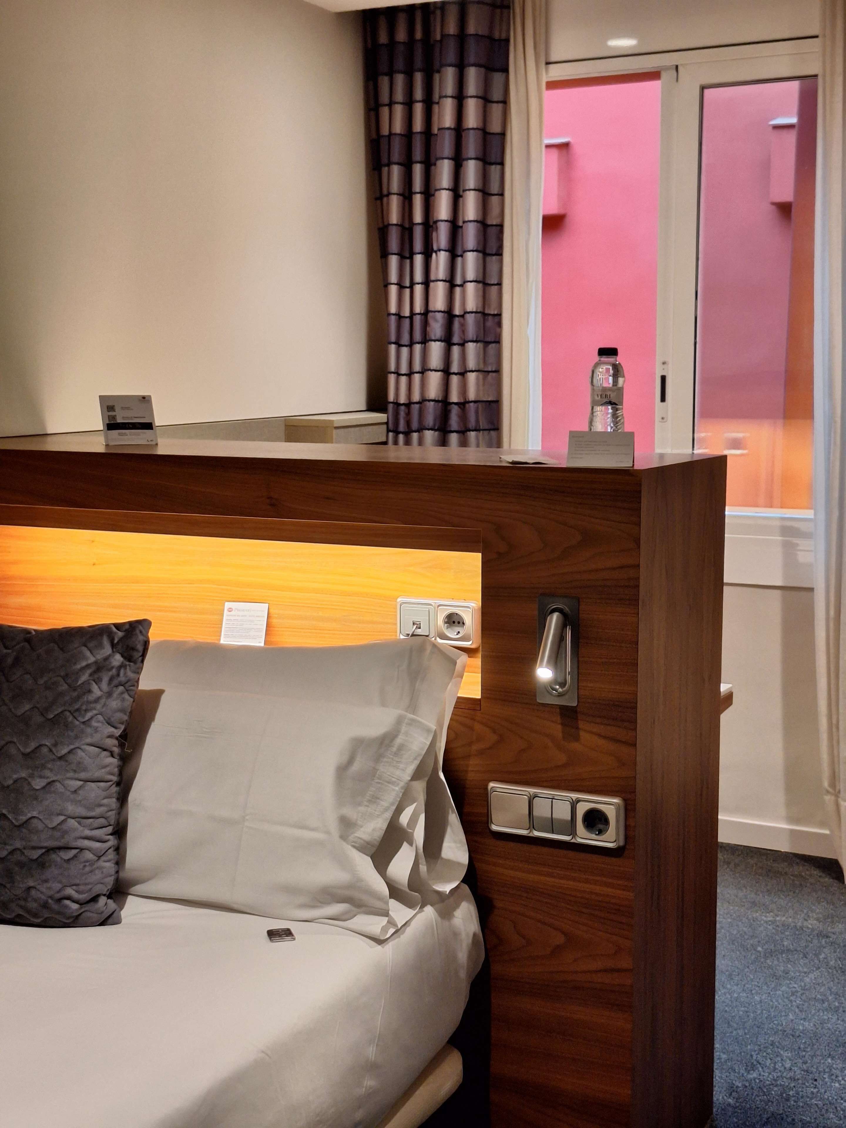 Habitación de hotel con cabecera de madera, cama con almohada gris, mesita de noche con una botella y vista a un edificio rosa a través de una ventana.