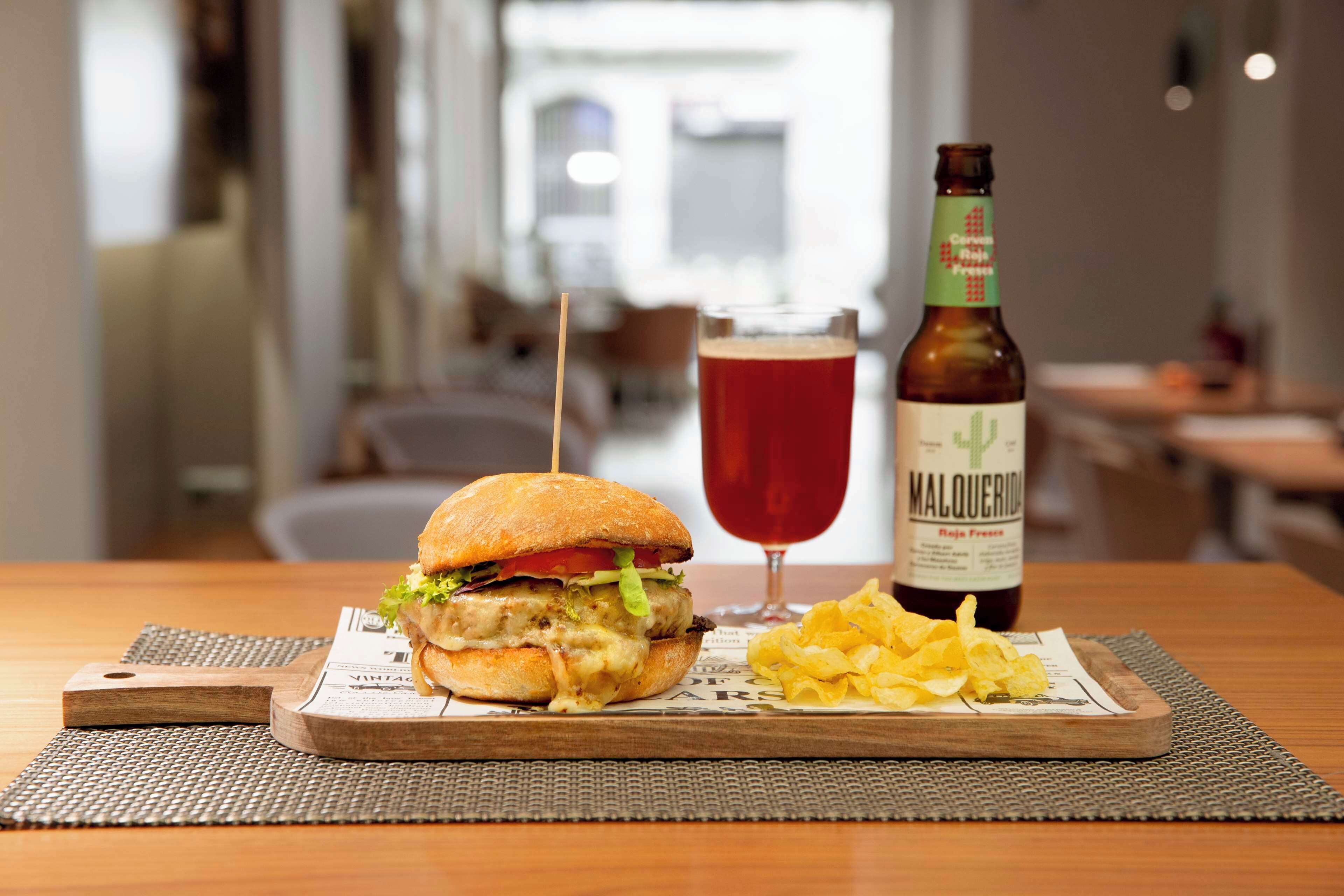 A meal with a burger, chips, and a bottle of Maluquer beer on a wooden table.