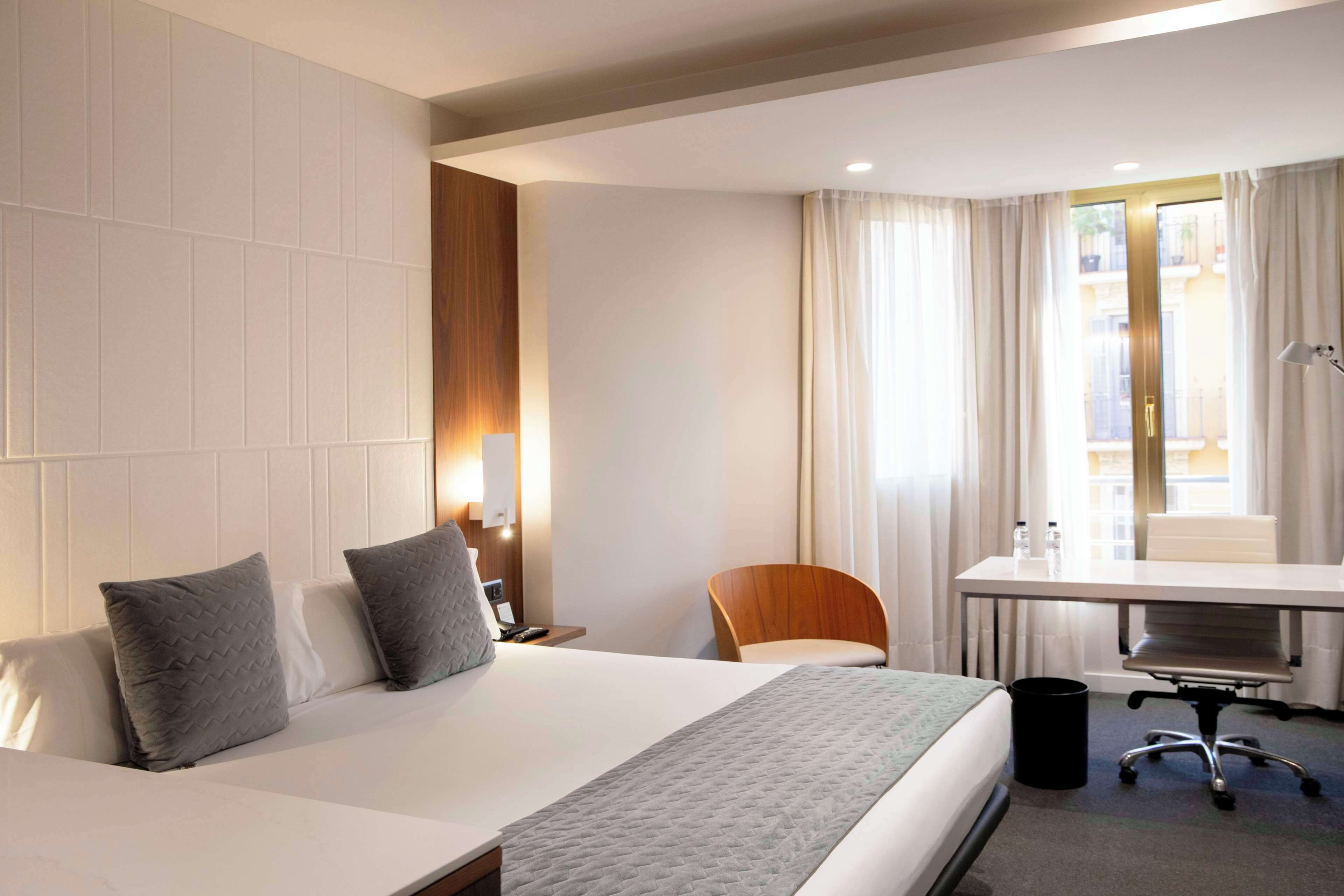 Ett hotellrum med en stor säng, ett skrivbord med en stol, ett litet bord med flaskor och ett fönster med gardiner.