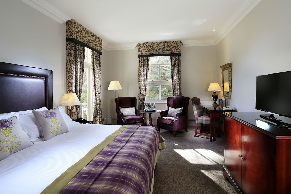 A hotel room with a bed, two armchairs, a desk with a TV, and a window with curtains.
