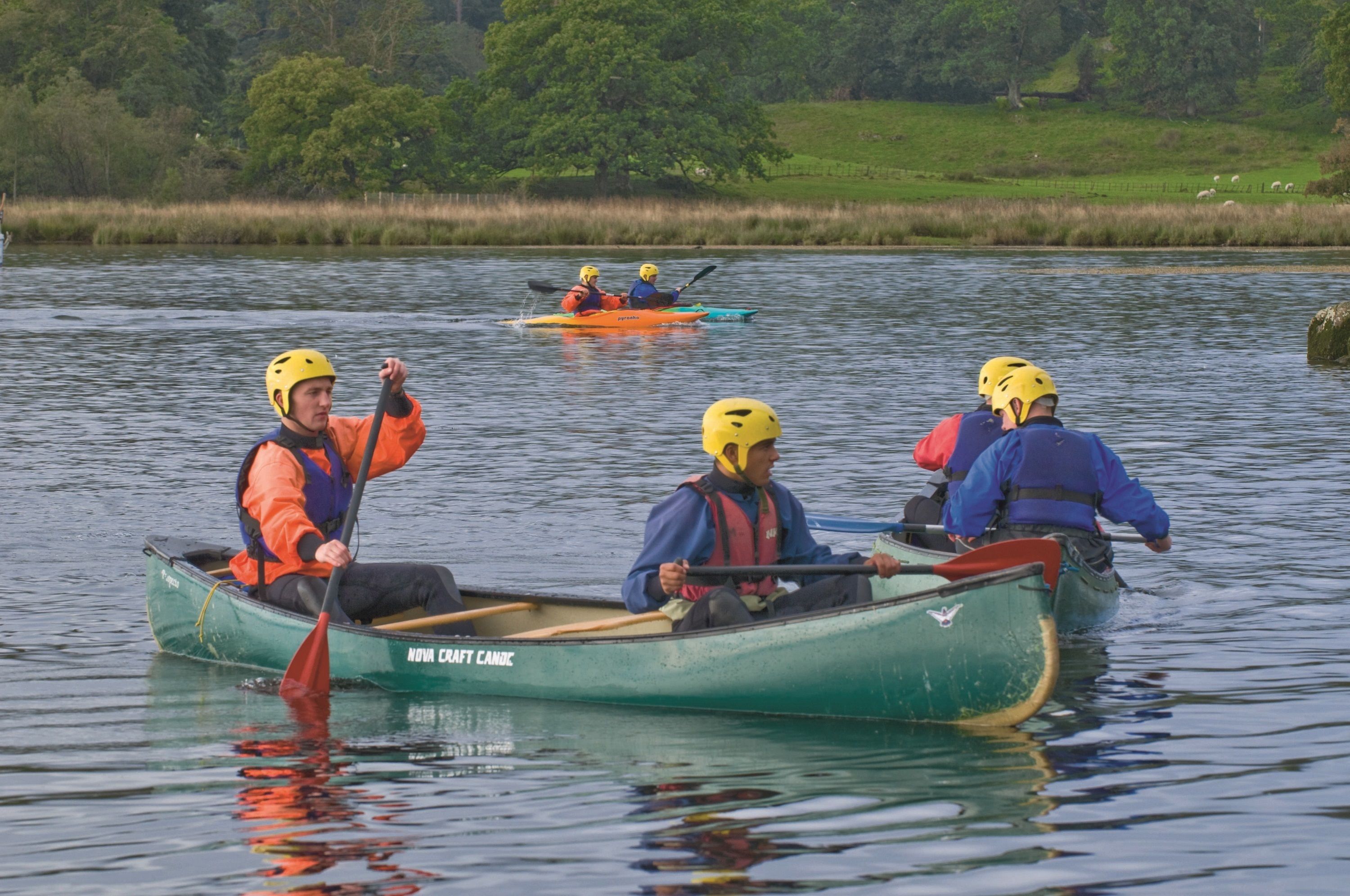 A group of people in a green canoe on a calm lake with grassy banks and trees in the background.