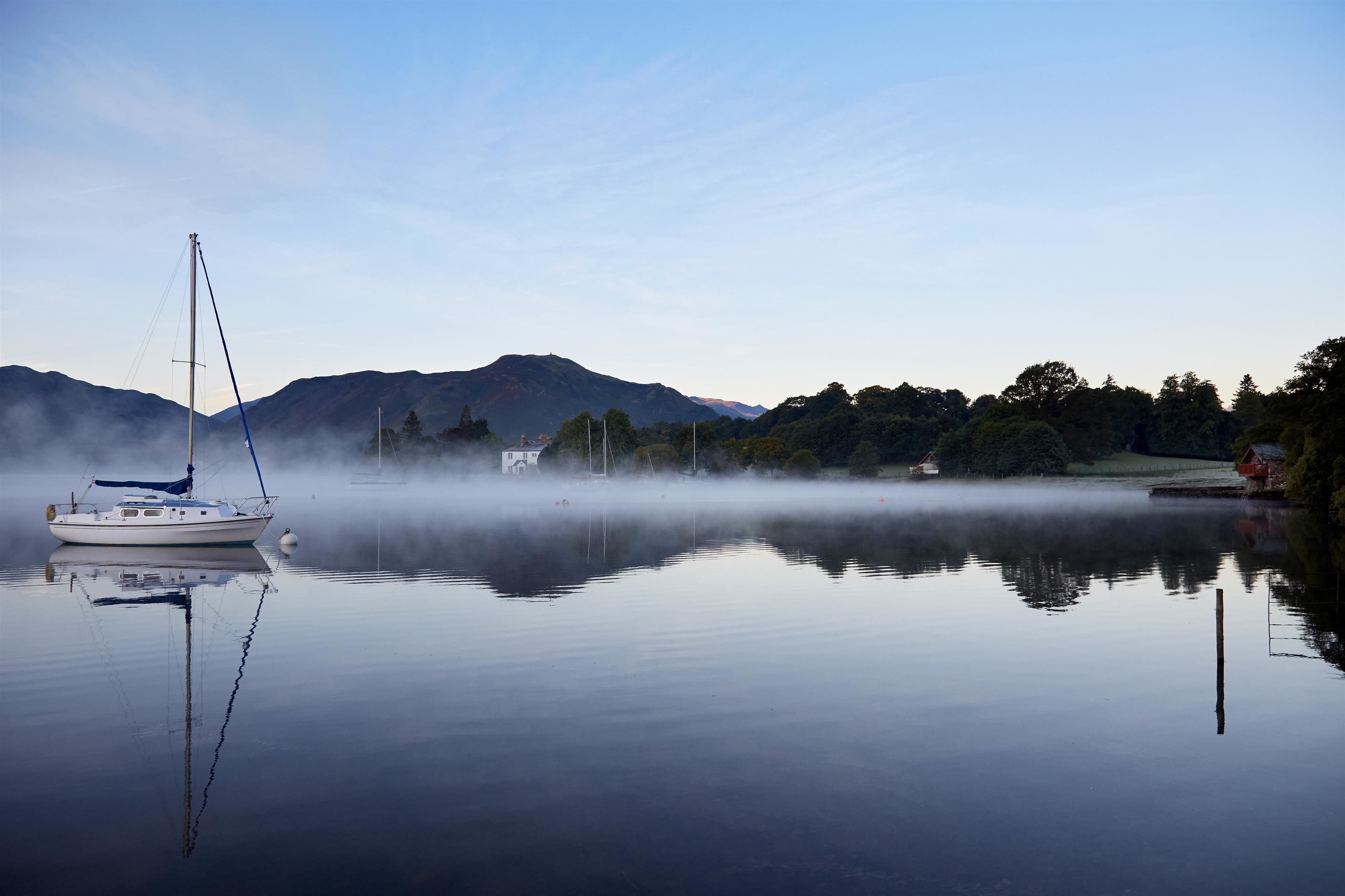 A calm lake with a sailboat anchored near the shore, surrounded by mist and mountains in the background.