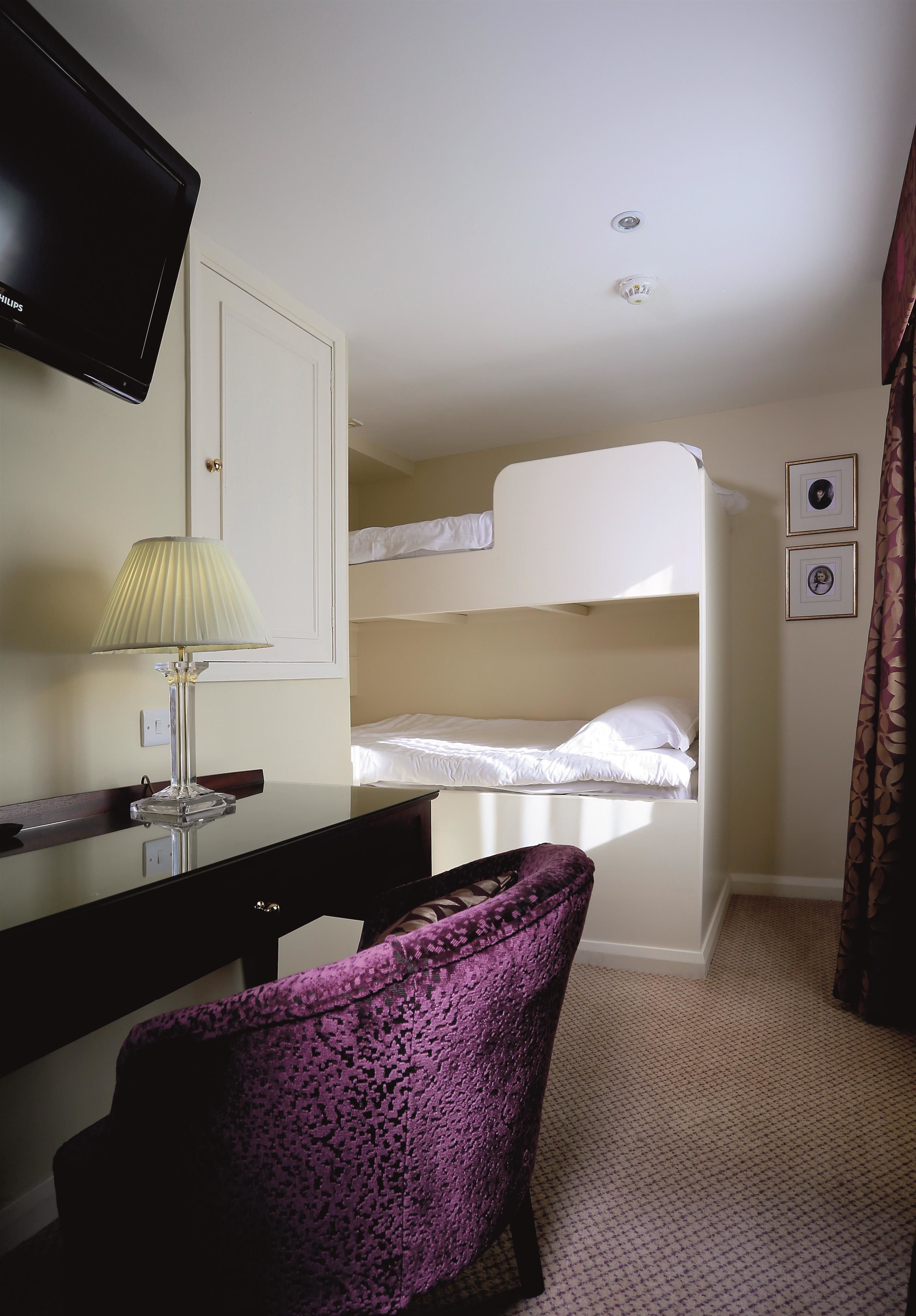 A hotel room with a bunk bed, a desk with a lamp, a chair, and a television on the wall.