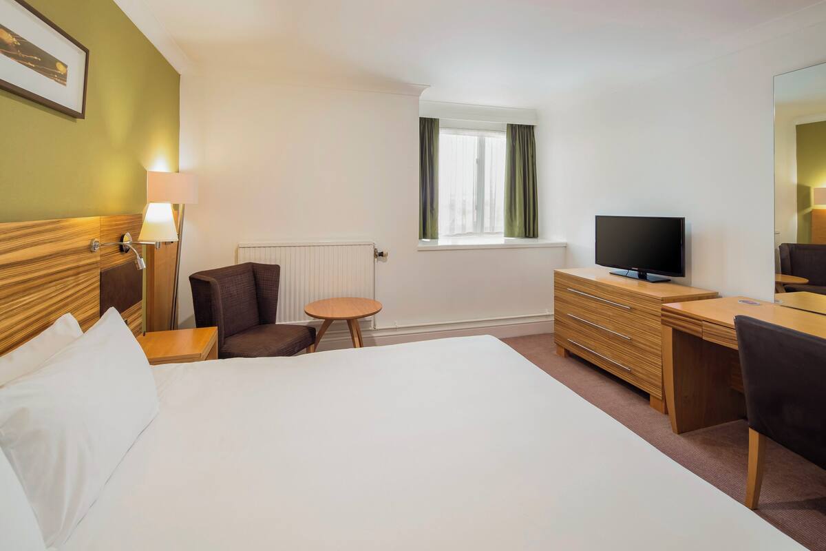Une chambre d’hôtel avec un grand lit, un bureau, une chaise, une télévision et une fenêtre avec des rideaux.