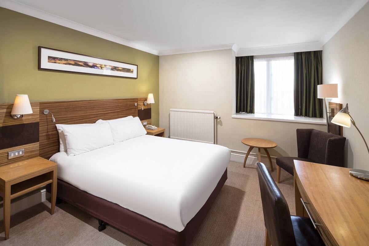Une chambre d’hôtel avec un grand lit, des tables de chevet, un bureau, une chaise, une fenêtre avec des rideaux et un tableau encadré au mur.