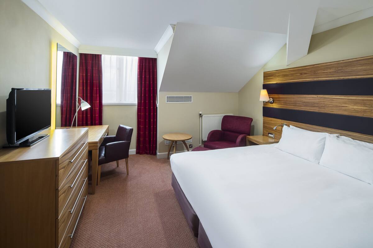 Une chambre d’hôtel avec un lit, un bureau avec une télévision, une chaise, une petite table et un rideau rouge.