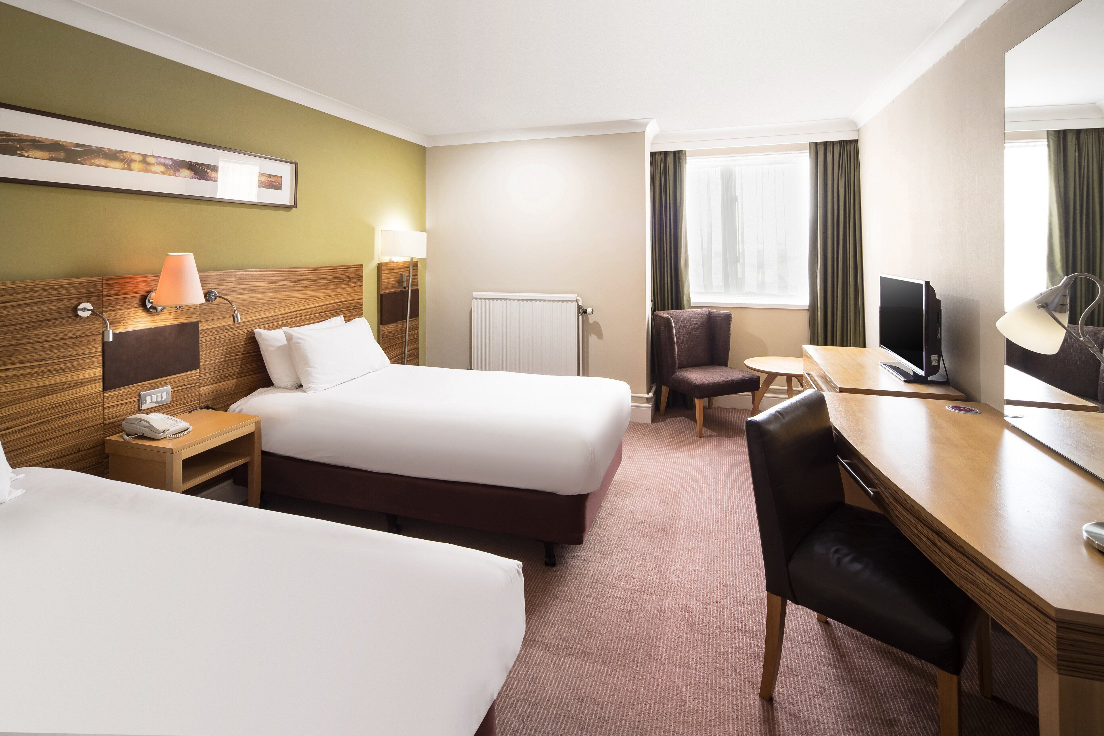A hotel room with two beds, a desk, a chair, a lamp, a TV, and a window with curtains.