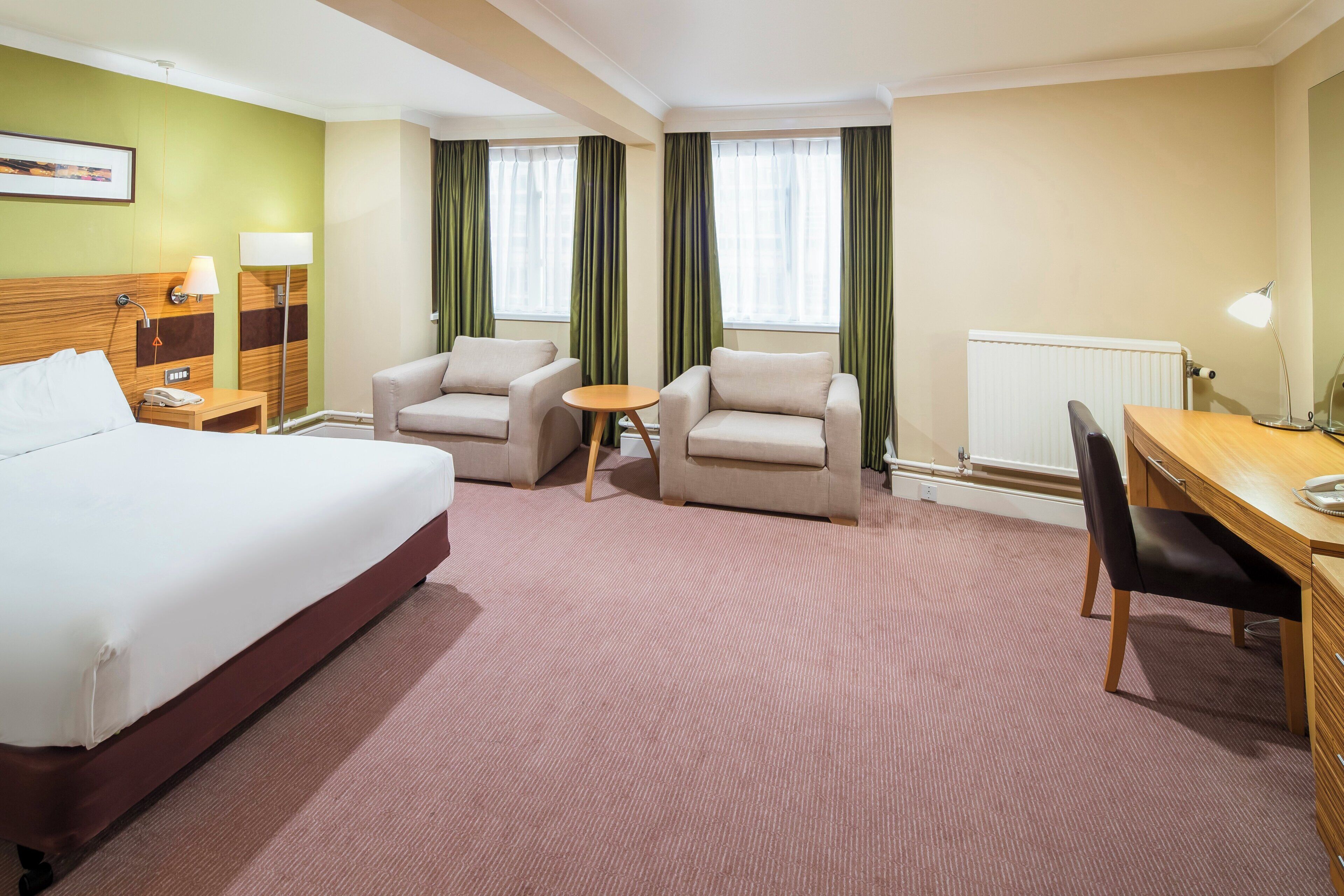 A hotel room with a bed, two armchairs, a small table, a desk, and a radiator.