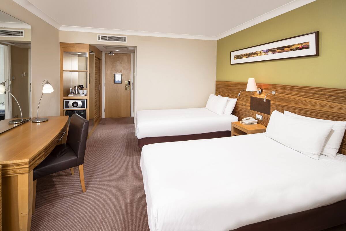 Une chambre d’hôtel avec deux lits, un bureau, une chaise, une lampe, un tél éphone et un tableau encadré au mur.