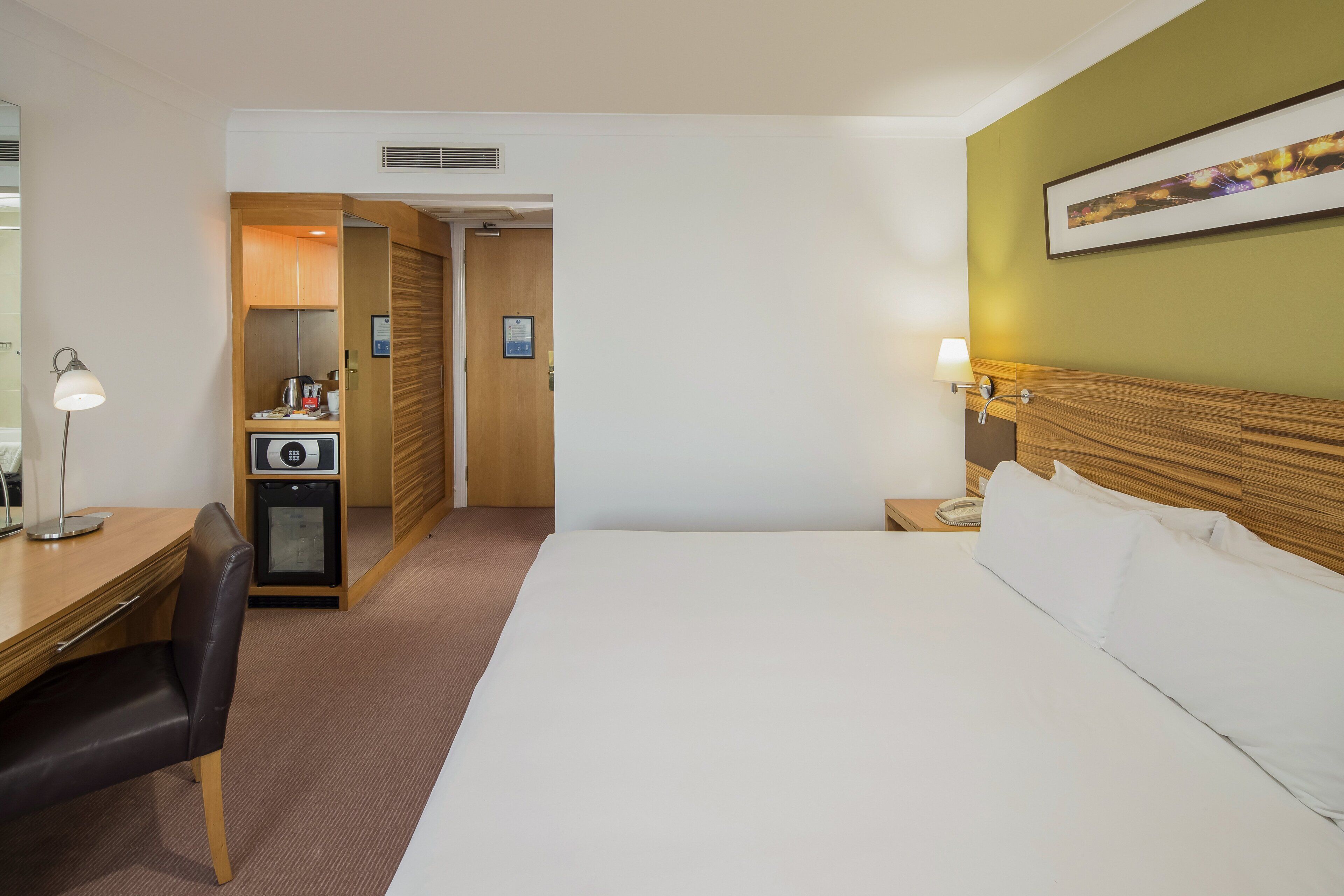A hotel room with a bed, a desk with a chair, a small kitchenette, and a bedside table with a lamp.