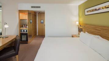 A hotel room with a bed, a desk with a chair, a small kitchenette, and a bedside table with a lamp.