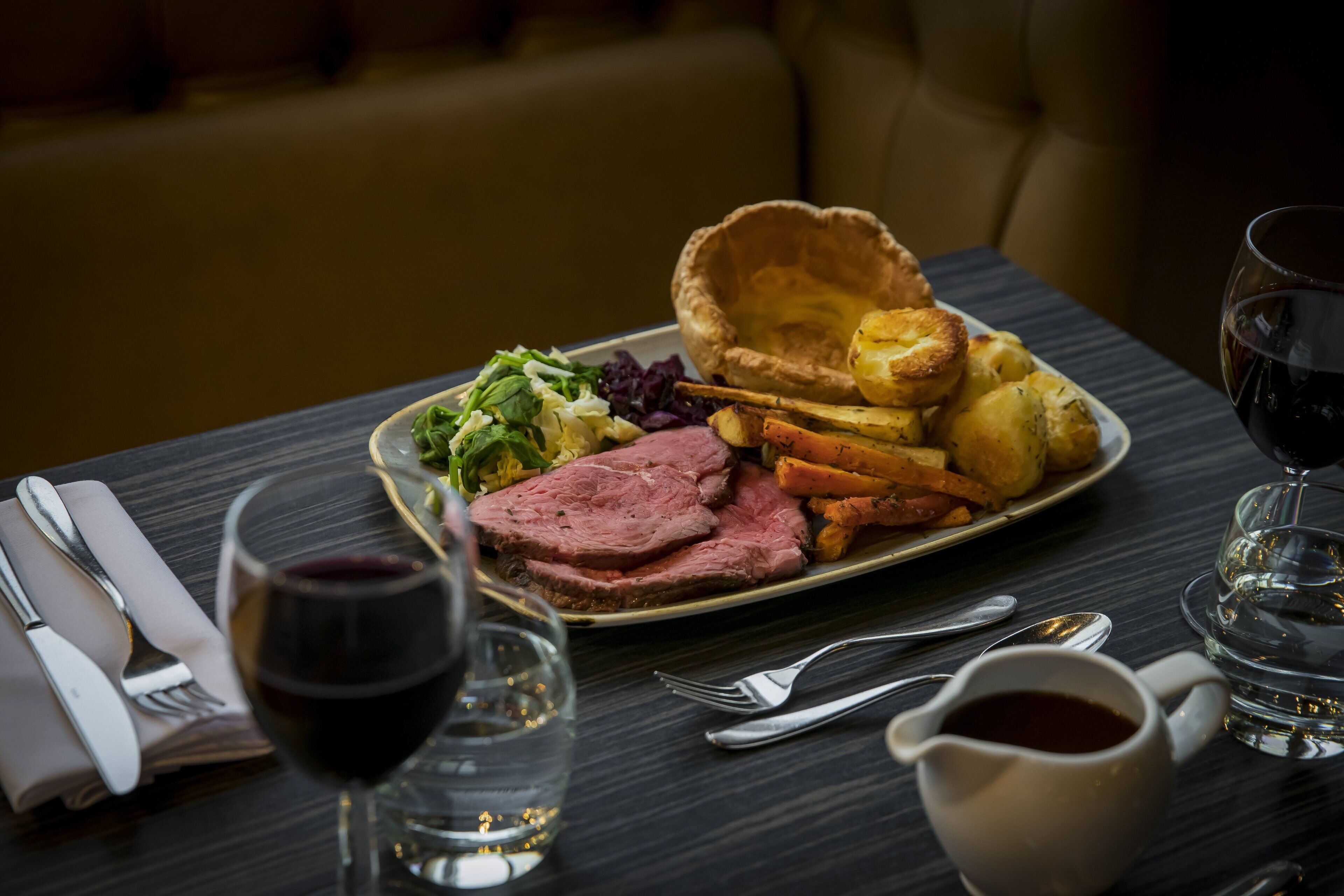 Een eettafel met een bord eten, waaronder gebraden rundvlees, Yorkshire pudding en groenten, plus een glas rode wijn en een kop thee.