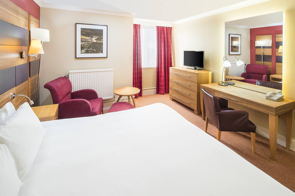 Une chambre d’hôtel avec un grand lit, un bureau avec une chaise, une petite table ronde, un fauteuil rouge, un canapé rouge, une télévision et un miroir.