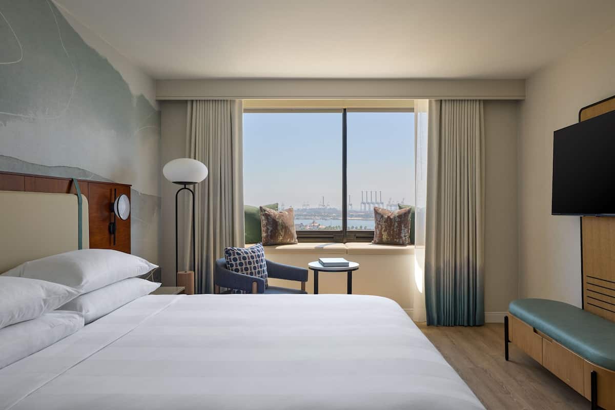 Une chambre d’hôtel avec un grand lit, une petite table, une chaise, un canapé, une télévision et une fenêtre donnant sur la ville.