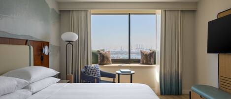 A hotel room with a large bed, a small table, a chair, a sofa, a TV, and a window with a city view.