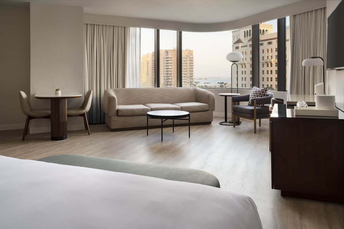 Une chambre d’hôtel moderne, dotée d’un grand lit, d’un canapé, d’une petite table et d’un bureau. La chambre dispose de grandes fenêtres offrant une vue sur de hauts immeubles et sur une étendue d’eau.