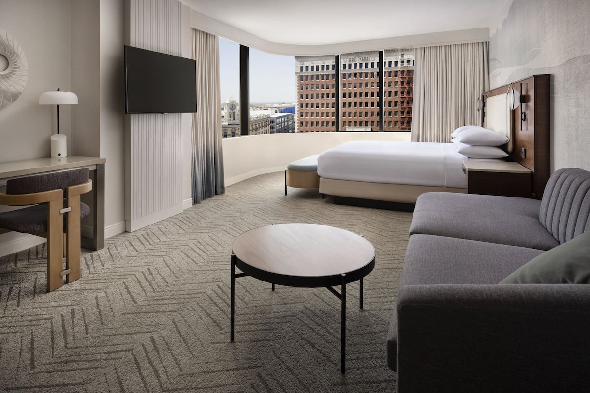 Une chambre d’hôtel avec un grand lit, un canapé, une petite table ronde, un bureau avec une lampe, un téléviseur fixé au mur et une vue sur le paysage urbain grâce à de grandes fenêtres.