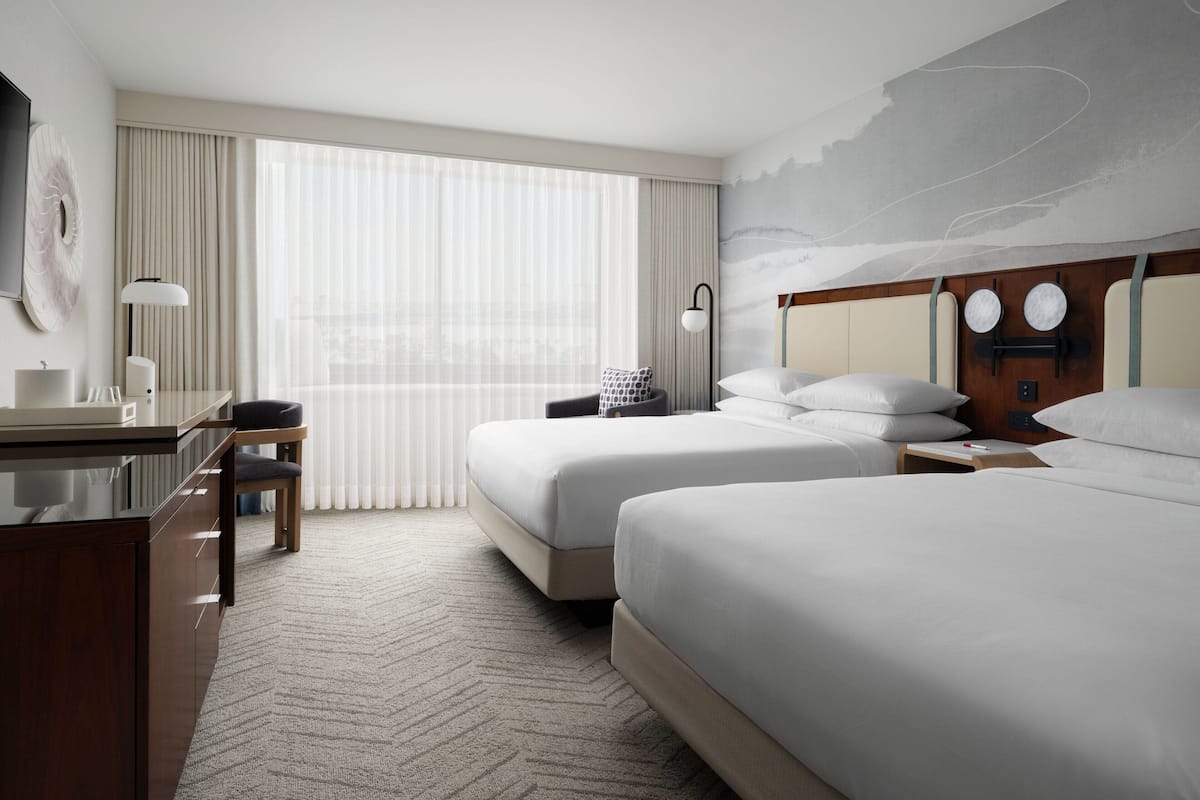 Une chambre d’hôtel avec deux lits, un bureau, une chaise, une lampe et une grande fenêtre avec des rideaux.