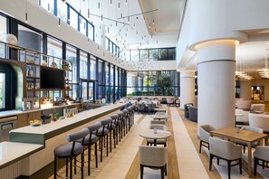 A modern bar area with high ceilings, large windows, and a variety of seating options including bar stools and tables.