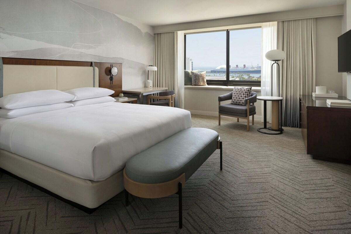Habitación de hotel con una cama grande, un escritorio, una silla, un banco, un televisor y una ventana con vistas a la ciudad.