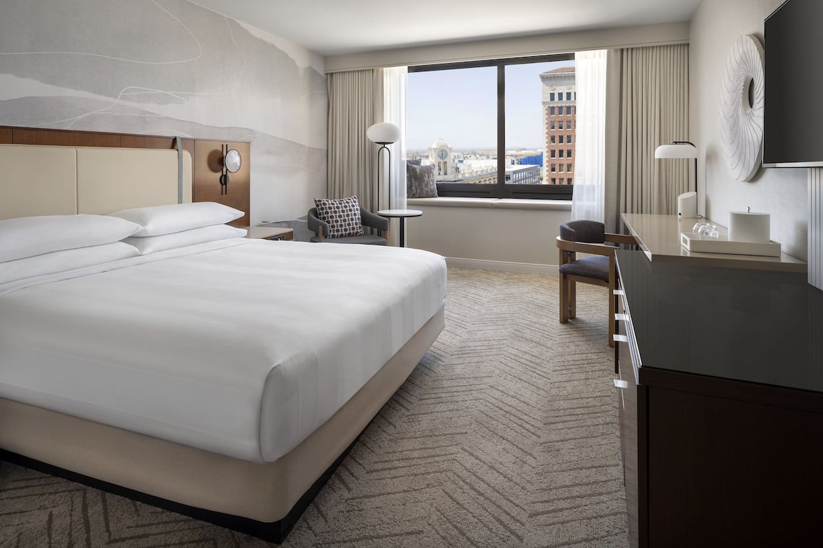 Une chambre d’hôtel avec un grand lit, un bureau, une chaise, une petite table et une vue sur la ville par la fenêtre.