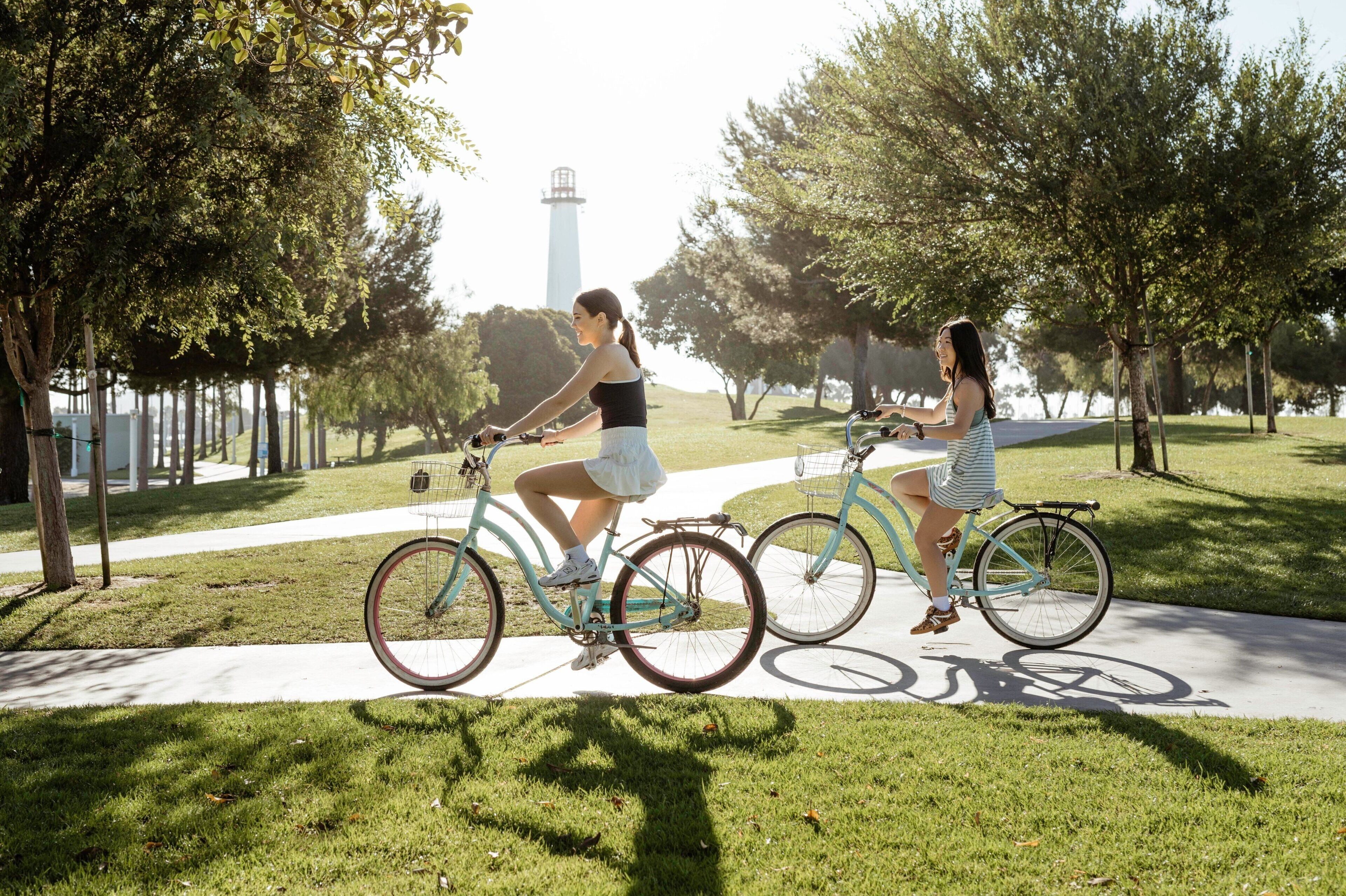 Two people riding bicycles on a paved path in a park with trees and a lighthouse in the background.