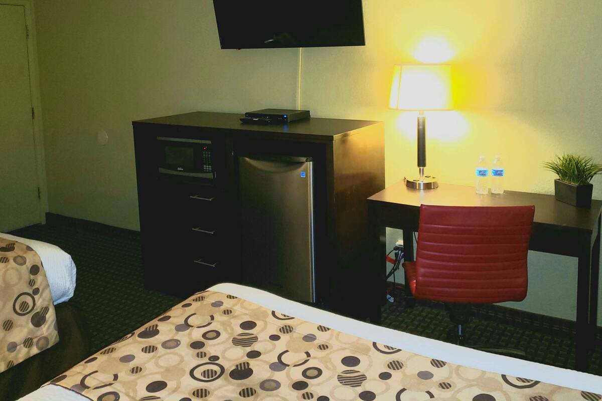A hotel room with a bed, a desk with a computer, a flat-screen TV, a refrigerator, a microwave, a lamp, and a potted plant.