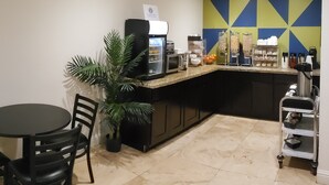 A modern kitchen with a coffee machine, a microwave, and a fruit bowl. There is a potted plant and a table with chairs.