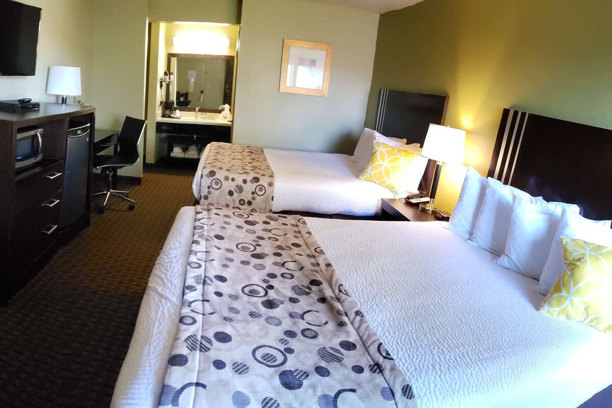 A hotel room with two beds, a desk with a chair, a TV, a mirror, and a bathroom visible through an open door.
