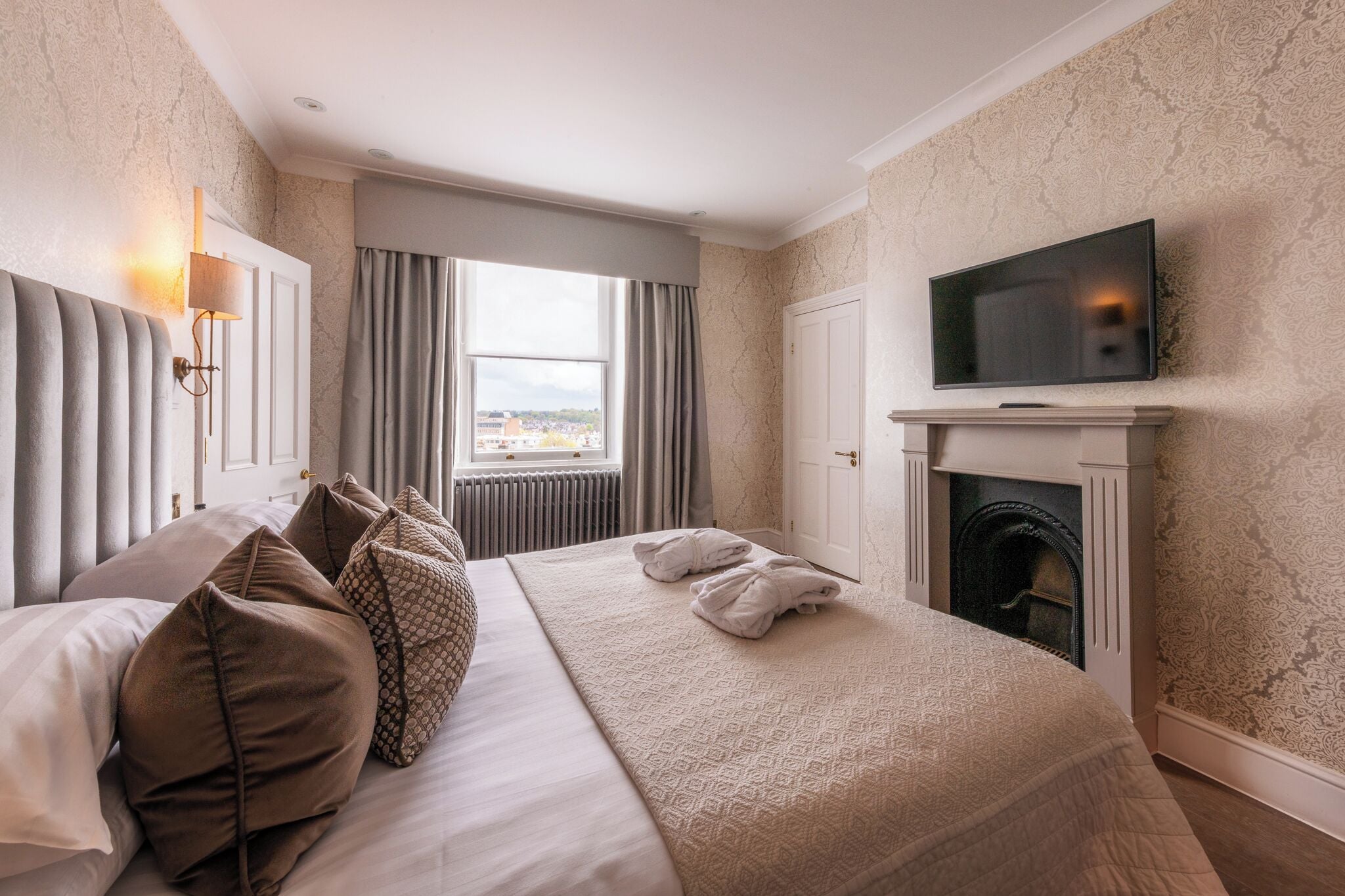 Een hotelkamer met een groot bed, een televisie boven een open haard, en uitzicht op de stad door het raam.