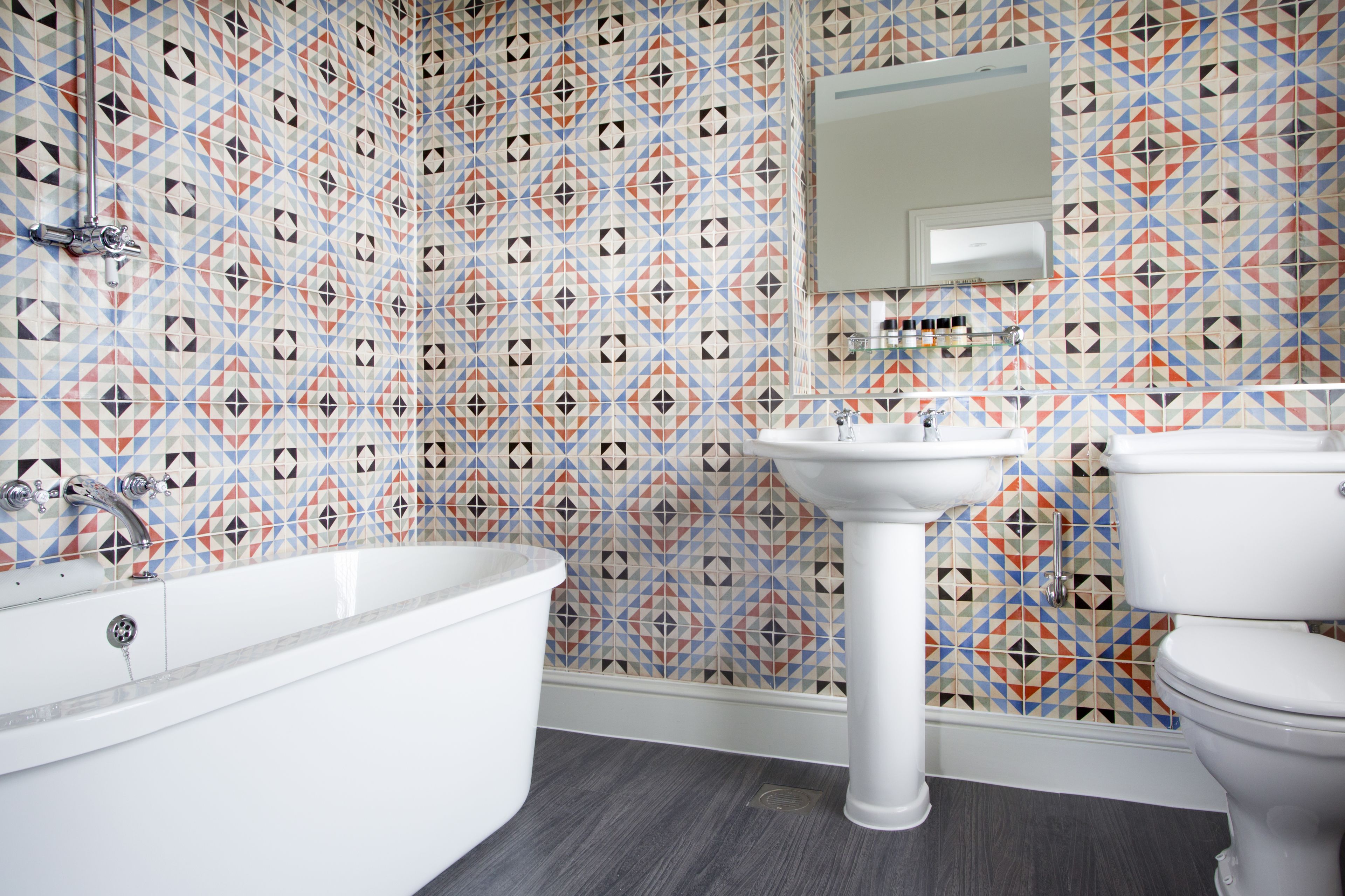 A bathroom with a geometric patterned tile wall, a white bathtub, a pedestal sink, and a toilet.