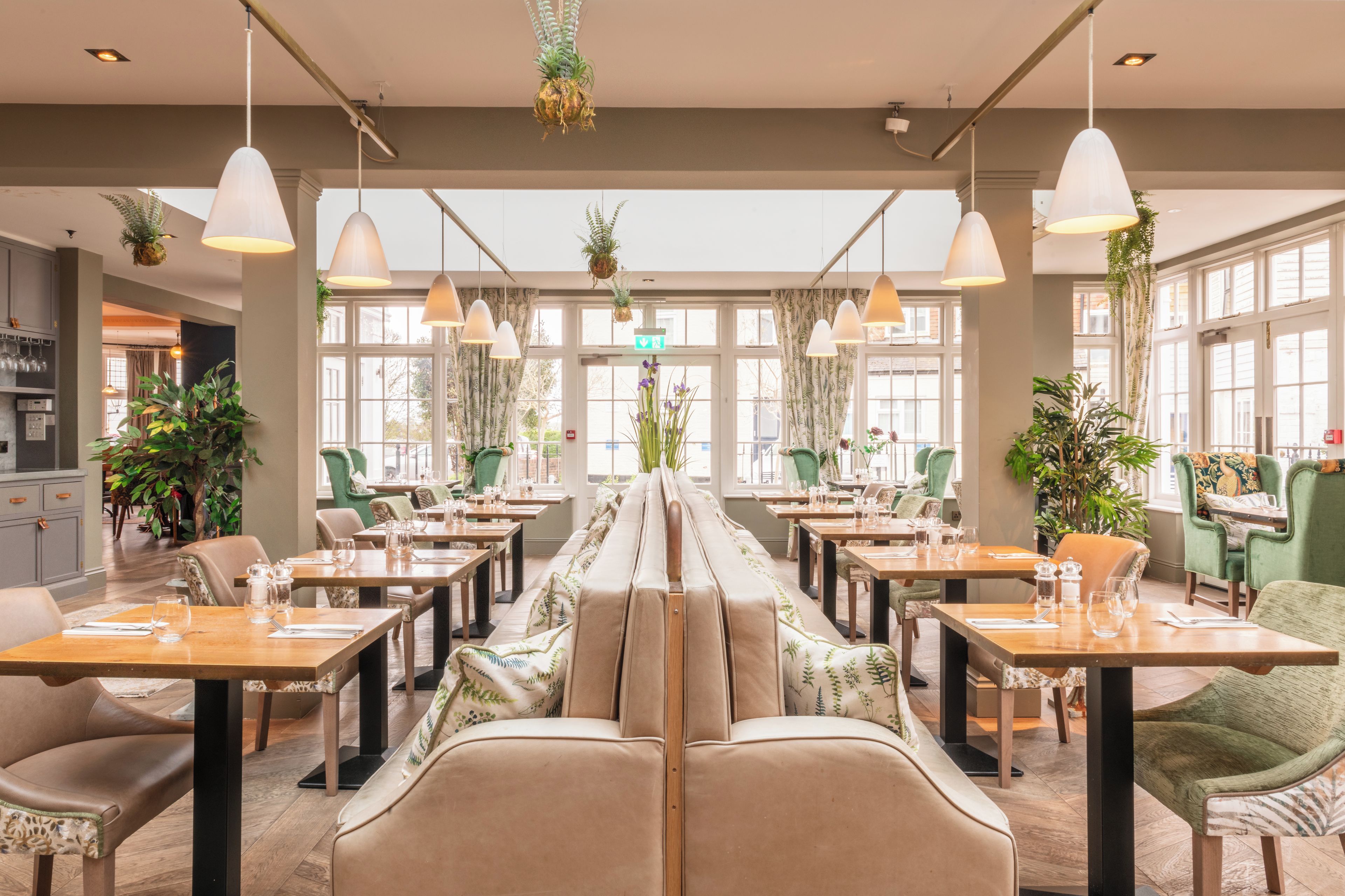 A restaurant with a long communal table, individual seating, hanging plants, and pendant lighting.