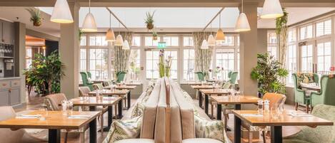 A restaurant with a long communal table, individual seating, hanging plants, and pendant lighting.